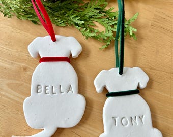 May include: Two white dog-shaped ornaments with red and green ribbon hangers. One ornament has the name "BELLA" and a red collar, the other has "TONY" and a green collar. The ornaments are on a wooden surface with greenery and a pinecone.