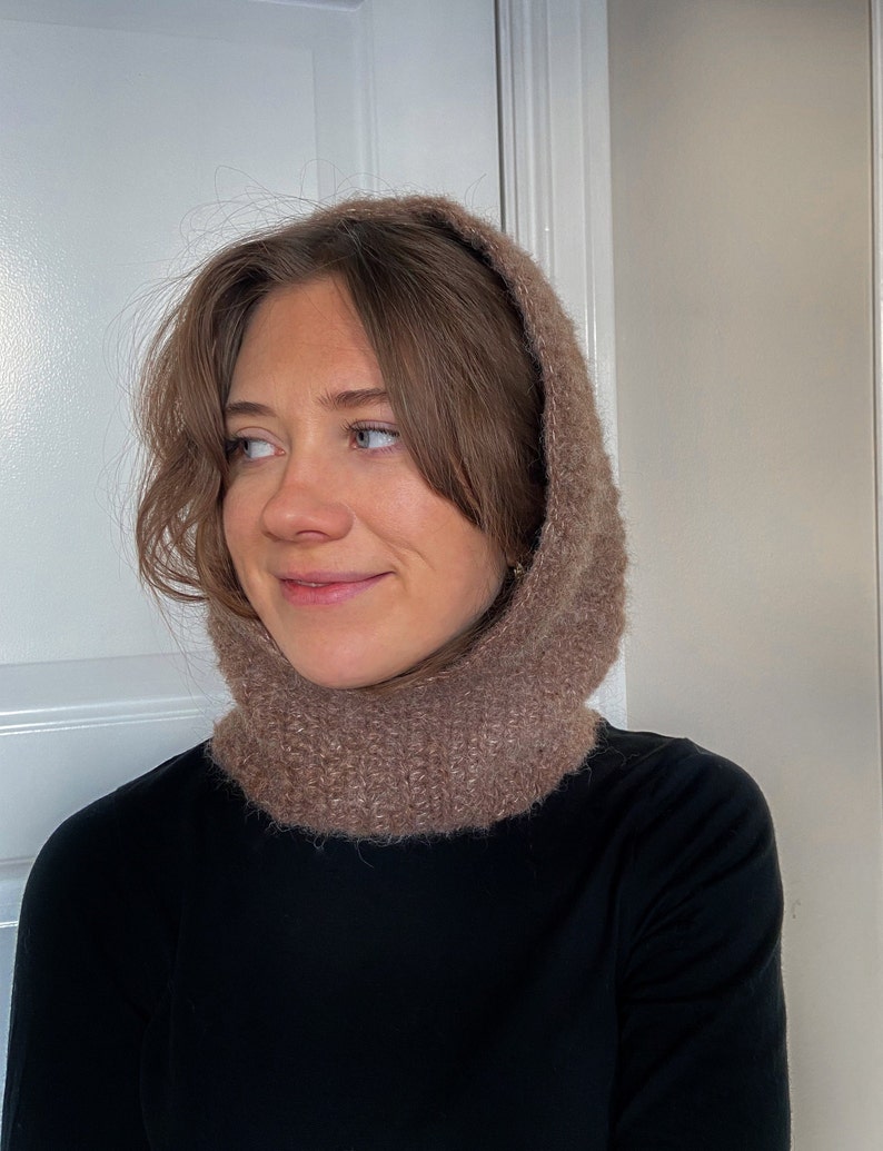 Op de afbeelding: Een bruine gebreide cowl neck met een capuchon. De cowl neck is gemaakt van een zacht, pluche garen.