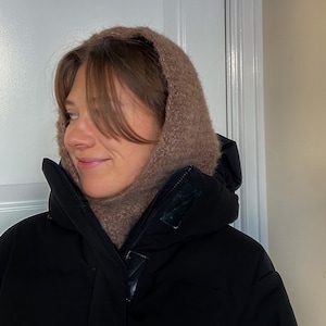 Op de afbeelding: Een vrouw draagt een zwarte winterjas met capuchon en een bruine, pluche sjaal. De capuchon is omhoog en de sjaal bedekt haar nek.