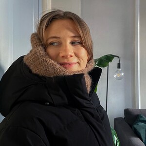 Op de afbeelding: Een vrouw draagt een zwarte pufferjas met een bruine gebreide sjaal om haar nek. Ze kijkt naar rechts van het frame.