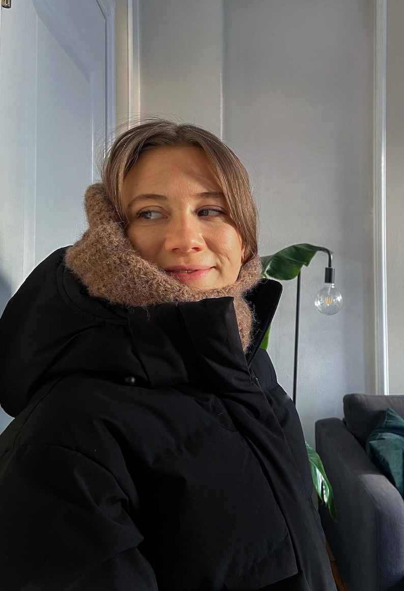 Op de afbeelding: Een vrouw draagt een zwarte pufferjas met een bruine gebreide sjaal om haar nek. Ze kijkt naar rechts van het frame.