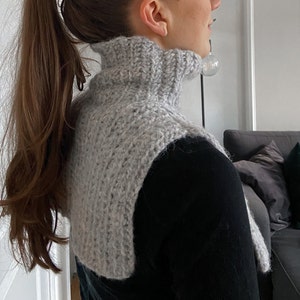May include: A grey knitted turtleneck collar with a textured pattern. The collar is worn over a black shirt.