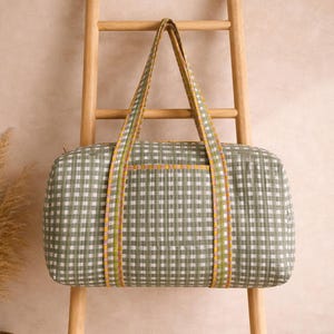 Könnte beinhalten: Eine grün-weiß karierte Reisetasche mit passenden Griffen und Zierleisten. Die Tasche ist rechteckig und hat eine kleine Tasche auf der Vorderseite. Die Griffe und Zierleisten weisen gelbe, orange und grüne Streifen auf. Die Tasche wird auf einer Holzleiter präsentiert.