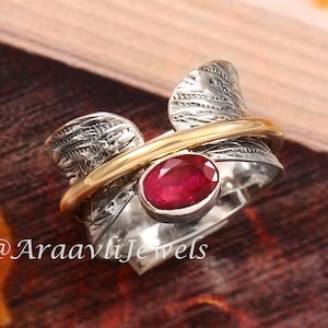 Könnte beinhalten: Ein silberner Ring mit einem goldenen Band und einem großen ovalen Rubin-Edelstein. Der Ring hat ein strukturiertes Design mit federartigen Details. Der Text "@AraavliJewels" ist sichtbar.