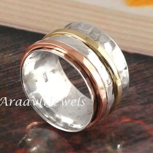 Könnte beinhalten: Ein silberner Spinner-Ring mit gehämmertem Muster und drei Bändern in Gold, Roségold und Silber. Der Ring ist ein Schmuckstück mit rotierendem Design. Der Ring liegt auf einer Holzoberfläche.
