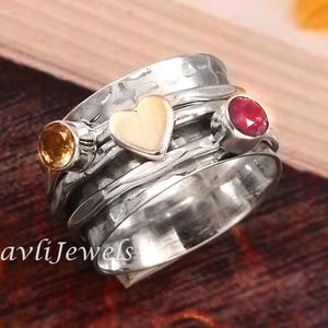 Könnte beinhalten: Ein silberner Spinner-Ring mit gehämmertem Muster. Er hat ein herzförmiges Detail und zwei Edelsteine: einen gelb-orangen Stein und einen roten Stein. Der Ring liegt auf einer Holzoberfläche.