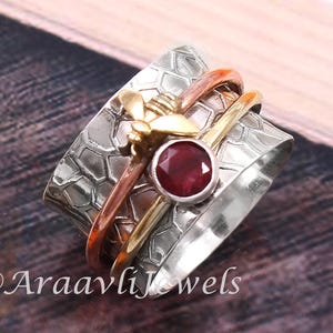 Puede incluir: Anillo giratorio plateado con una banda texturizada y una gema roja central. El anillo presenta una pequeña abeja dorada y dos bandas giratorias en tonos cobre y oro. El texto "@AraavliJewels" es visible.