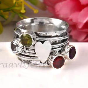 Könnte beinhalten: Ein silberner Spinner-Ring mit Herz-Akzent und mehreren Edelsteinen. Der Ring hat einen Peridot, einen klaren Edelstein, einen Granat und einen Amethyst. Der Ring liegt auf einer weißen Oberfläche mit Blumen im Hintergrund.