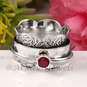 Könnte beinhalten: Ein silberfarbener Ring mit einem strukturierten Band und einem zentralen, runden, roten Edelstein. Der Ring hat ein mehrschichtiges Design mit einem dekorativen, gemusterten Innenband. Der Ring ist vor einem Hintergrund aus Blumen platziert.