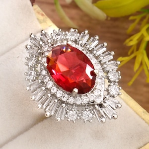 May include: A silver ring with a large, oval-cut, red gemstone at its centre, encircled by smaller, clear, baguette-cut and round gemstones. The ring is set in a silver band and is presented in a cream-coloured ring box.