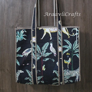 Könnte beinhalten: Schwarze Tragetasche mit Dschungel-Print mit Zebras, Leoparden und Papageien. Die Tasche hat ein gestepptes Design mit gestreiftem Besatz und Metallgriffen. Der Text "AraavliCrafts" ist auf der Tasche aufgedruckt.