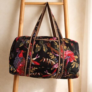 Könnte beinhalten: Schwarze, gesteppte Reisetasche mit einem lebendigen Blumen- und Vogelmuster in Rot, Lila und Grün. Die Tasche hat zwei Griffe und wird auf einer Holzleiter präsentiert. Das Design der Tasche umfasst botanische und Vogel-Elemente.