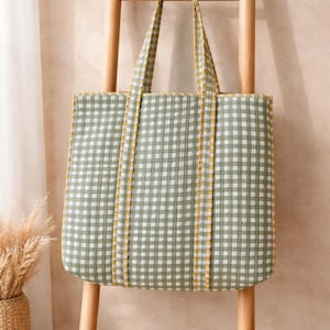 Peut inclure: Un sac fourre-tout à carreaux verts et blancs avec une bordure jaune, présenté sur un support en forme d'échelle en bois. Le sac a des poignées assorties et un design simple. L'arrière-plan présente un mur neutre et un panier en osier.