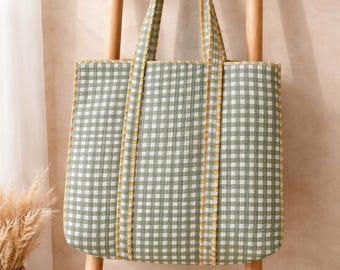 Puede incluir: Un bolso tote a cuadros verdes y blancos con ribete amarillo, exhibido en un soporte de escalera de madera. El bolso tiene asas a juego y un diseño sencillo. El fondo presenta una pared neutra y una cesta de mimbre.