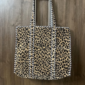 Könnte beinhalten: Eine Tragetasche mit Leopardenmuster und schwarz-weiß gestreiften Verzierungen und Griffen. Die Tasche hat einen beigefarbenen Hintergrund mit schwarzen Flecken, und die Griffe haben eine silberne Metallstange.