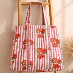 Könnte beinhalten: Gesteppte Tragetasche mit weiß-rosa Streifenmuster und Erdbeermotiv. Die Tasche hat zwei Henkel und hängt an einer Holzleiter. Die Tasche ist etwa 38 cm breit und 41 cm hoch.