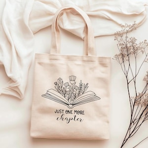 Könnte beinhalten: Eine beige Canvas-Tragetasche mit einer schwarzen Umrandung eines offenen Buches, aus dem Blumen wachsen. Der Text "Just one more chapter" ist unter dem Buch gedruckt.