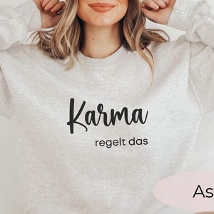 May include: A gray sweatshirt with the text "Karma regelt das" in black lettering. The sweatshirt is shown on a model and the color of the sweatshirt is labeled "Ash".