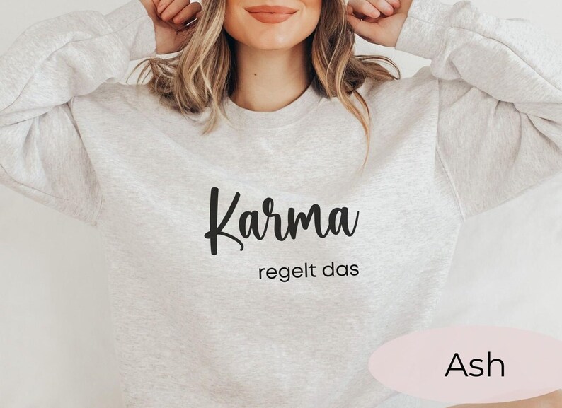 May include: A gray sweatshirt with the text "Karma regelt das" in black lettering. The sweatshirt is shown on a model and the color of the sweatshirt is labeled "Ash".