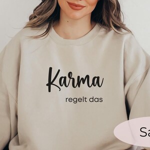 May include: A beige sweatshirt with the text "Karma regelt das" in black lettering. The sweatshirt is on a white surface. The color of the sweatshirt is "Sand".