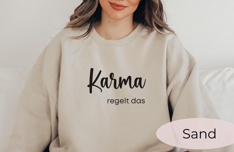 May include: A beige sweatshirt with the text "Karma regelt das" in black lettering. The sweatshirt is on a white surface. The color of the sweatshirt is "Sand".
