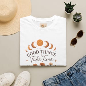 Könnte beinhalten: Ein weißes T-Shirt mit einer Grafik der Mondphasen in Orange- und Brauntönen, dem Schriftzug "GOOD THINGS Take Time" in Dunkelbraun sowie kleinen Sternsymbolen und orangefarbenen Blumen am unteren Rand des Designs.