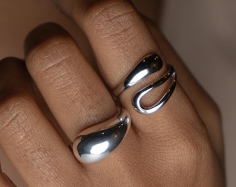 May include: Two silver rings on a hand. The ring on the index finger is a simple, smooth, oval shape. The ring on the middle finger is a more abstract, open design.