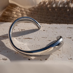 Puede incluir: Un brazalete de plata con un dise&ntilde;o curvo y liso. El brazalete est&aacute; hecho de metal y tiene un acabado pulido.