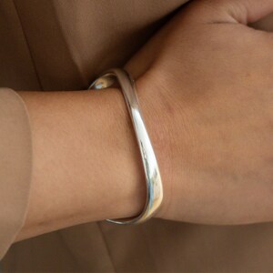 Puede incluir: Un brazalete de plata con un dise&ntilde;o curvo y abierto.