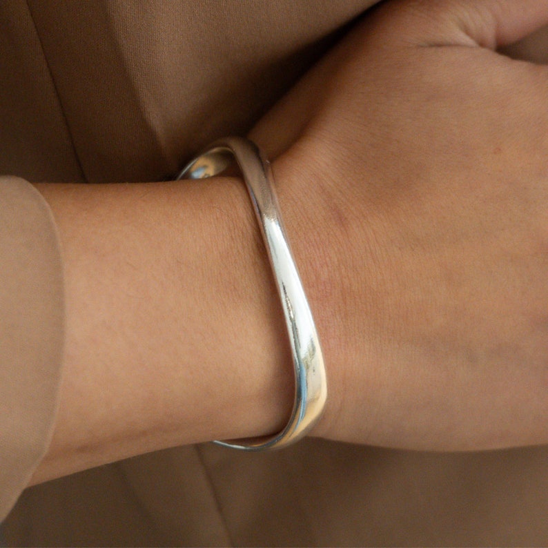 Puede incluir: Un brazalete de plata con un dise&ntilde;o curvo y abierto.