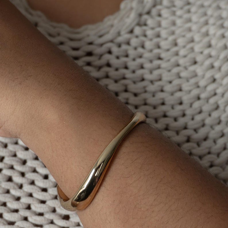 Puede incluir: Un brazalete curvo de color dorado. El brazalete es liso y tiene un dise&ntilde;o simple y moderno.