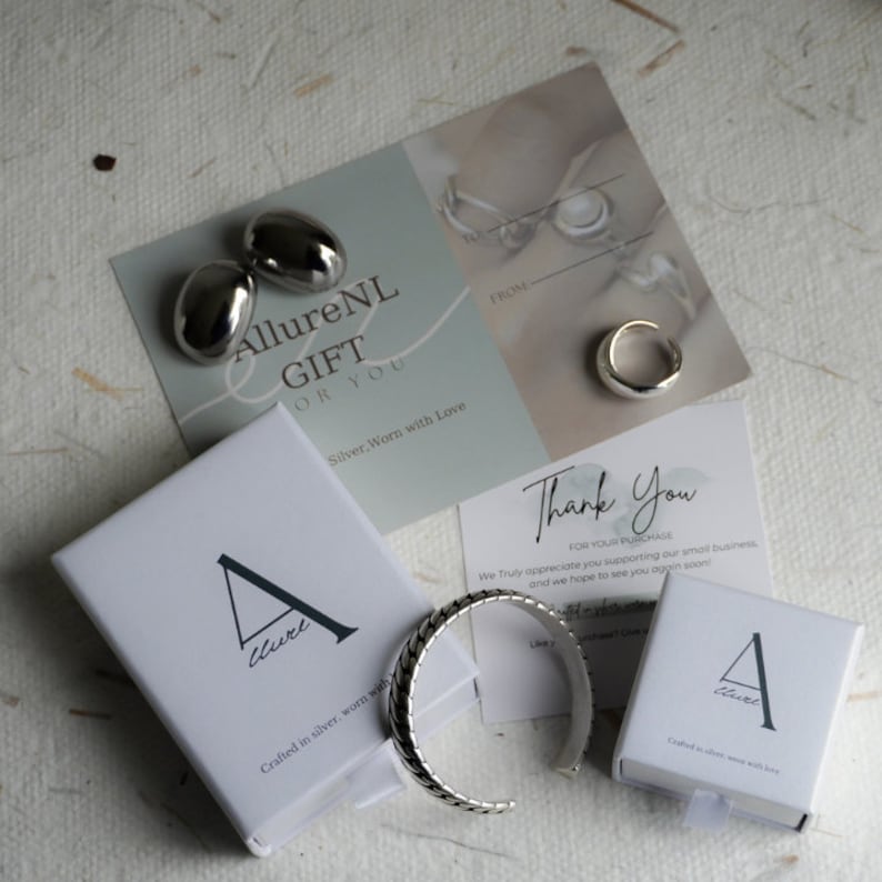 Puede incluir: Dos cajas plateadas con el texto "AllureNL GIFT OR YOU" y "Crafted in silver, worn with love" en la parte delantera. Las cajas contienen joyas de plata, incluyendo un par de pendientes, un anillo y una pulsera.