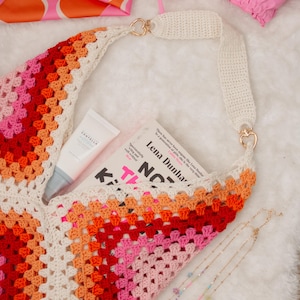 May include: A colorful, handmade crochet bag with a white strap and gold-colored heart-shaped hardware. The bag is filled with a book, skincare product, and other accessories. A pink bikini top and a patterned scarf are also visible.