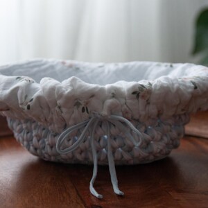 Könnte beinhalten: Handgefertigter, ovaler Korb mit hellblau gehäkeltem Äußeren und einem Stofffutter mit Blumenmuster. Der Korb hat einen hellblauen Kordelverschluss. Ideal für Heimdekoration und Aufbewahrung.