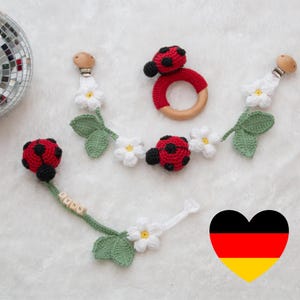 Könnte beinhalten: Ein gehäkeltes Marienkäfer-Babyspielzeug mit einem Holzring, weißen Blüten und grünen Blättern. Das Spielzeug ist an einem Holzclip befestigt, an dem der Name "Lynn" in Holzbuchstaben geschrieben ist.