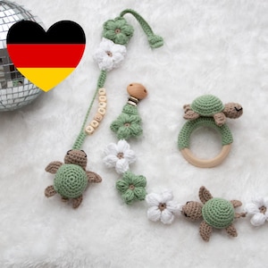 Könnte beinhalten: Ein grünes und weißes gehäkeltes Babyspielzeug mit einem Holzring und einem Schnullerclip. Das Spielzeug besteht aus einer Kette von Blumen und Schildkröten. Der Schnullerclip trägt den Namen "Valerie" darauf.
