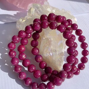 May include: Two strands of deep pink gemstone beads, likely ruby, are arranged in a circle on a white surface. The beads are smooth and round, and they are clustered together in a close-up view.