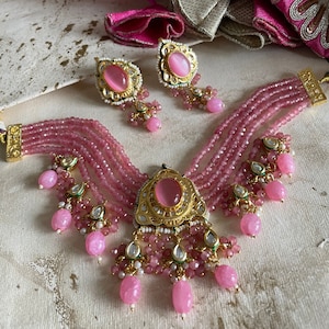 May include: A pink and gold layered beaded necklace with a large pink stone pendant and matching earrings. The necklace has multiple strands of pink beads and a gold pendant with a pink stone in the center. The earrings are also pink and gold with a pink stone in the center.
