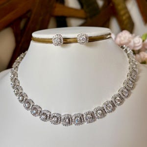 Könnte beinhalten: Ein silberfarbenes Halsketten- und Ohrstecker-Set. Die Halskette und die Ohrringe sind mit quadratischen, klaren Edelsteinen besetzt. Der Schmuck wird auf einer weißen Büste präsentiert, mit einem verschwommenen Hintergrund aus Holzmöbeln und Blumen.