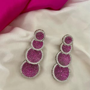 Può includere: Un paio di orecchini d'argento con pietre rosa. Ogni orecchino ha quattro pietre rotonde, che diminuiscono di dimensione dal basso verso l'alto.