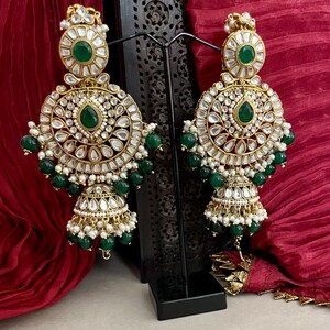 May include: A pair of gold-tone earrings with green and white stones. The earrings are dangling and have a round shape with a smaller round shape at the bottom. The earrings are on a black stand.