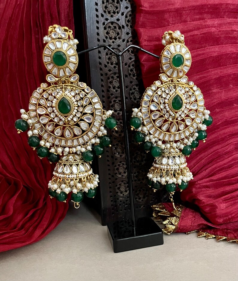 May include: A pair of gold-tone earrings with green and white stones. The earrings are dangling and have a round shape with a smaller round shape at the bottom. The earrings are on a black stand.