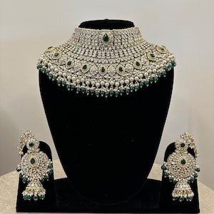 May include: A traditional Indian bridal necklace set with a large, ornate choker and matching earrings. The set is made of gold-toned metal and features white pearls, clear rhinestones, and emerald green stones.