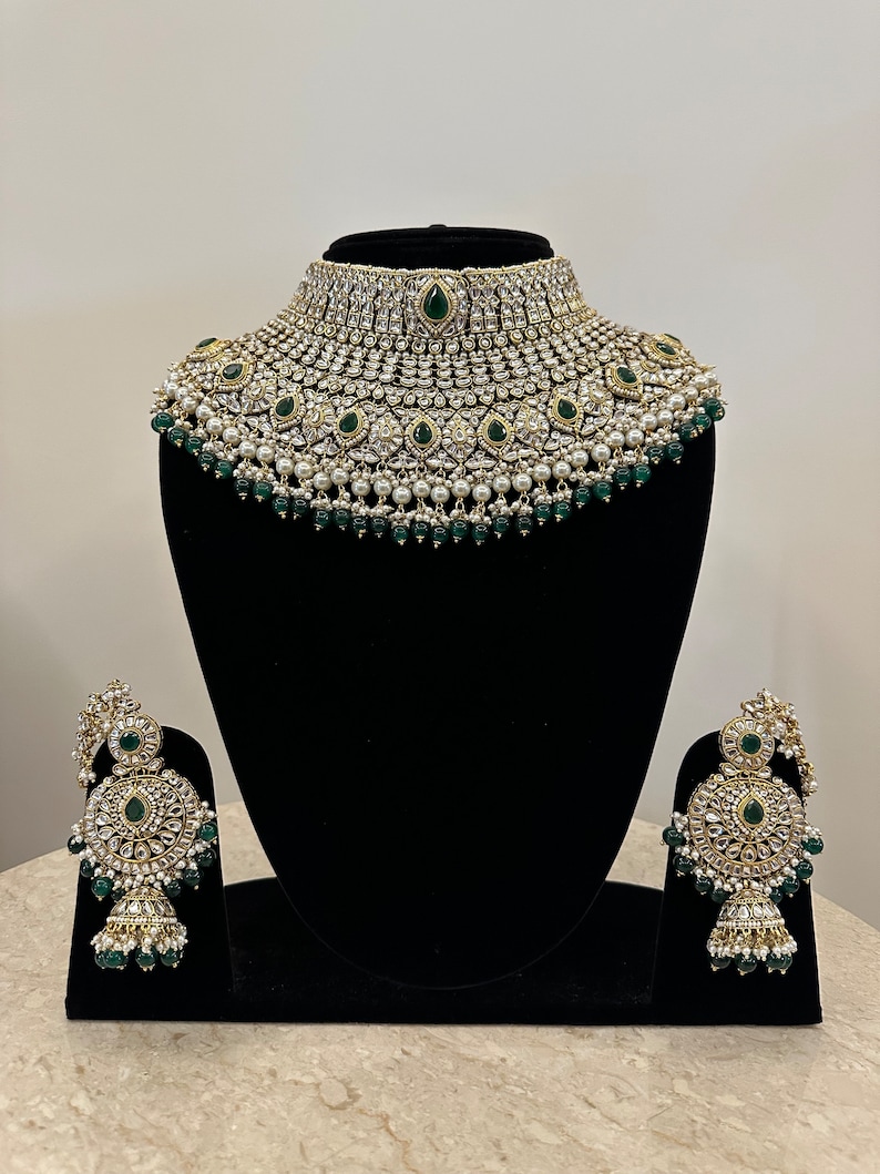 May include: A traditional Indian bridal necklace set with a large, ornate choker and matching earrings. The set is made of gold-toned metal and features white pearls, clear rhinestones, and emerald green stones.