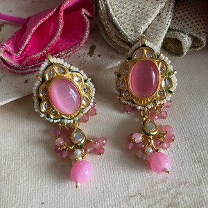 May include: A pair of pink and gold earrings with a pink stone in the center. The earrings have a dangling design with pink and white beads.