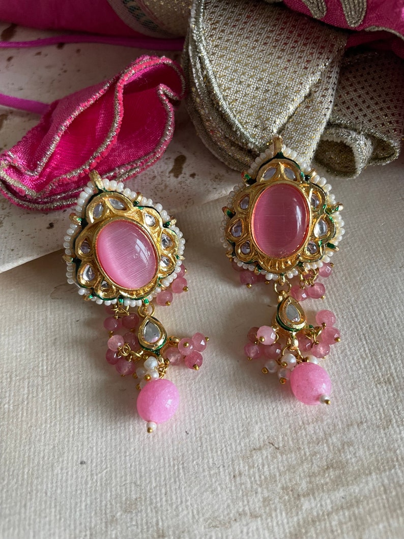 May include: A pair of pink and gold earrings with a pink stone in the center. The earrings have a dangling design with pink and white beads.