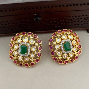 May include: A pair of gold-toned stud earrings. Each earring features a square, emerald-green center stone surrounded by a halo of small, clear stones. The outer edge is adorned with ruby-colored stones.