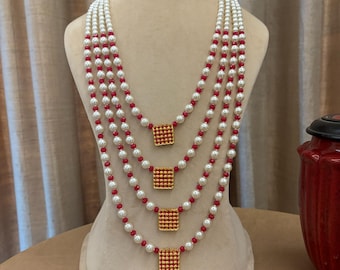 May include: A multi-strand necklace with white pearls and red beads, interspersed with square gold-colored pendants. The necklace is displayed on a mannequin, with loose gold beads scattered on the surface.