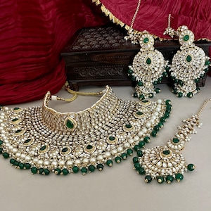 May include: A traditional Indian bridal jewelry set featuring a large, ornate necklace with green and white stones, matching earrings, and a headpiece. The set is made of gold and silver and is decorated with pearls and beads.