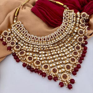 May include: A gold-toned statement necklace with intricate detailing. The necklace features a layered design with red and white accents, including small pearls and red beads. The necklace is displayed on a neutral background.